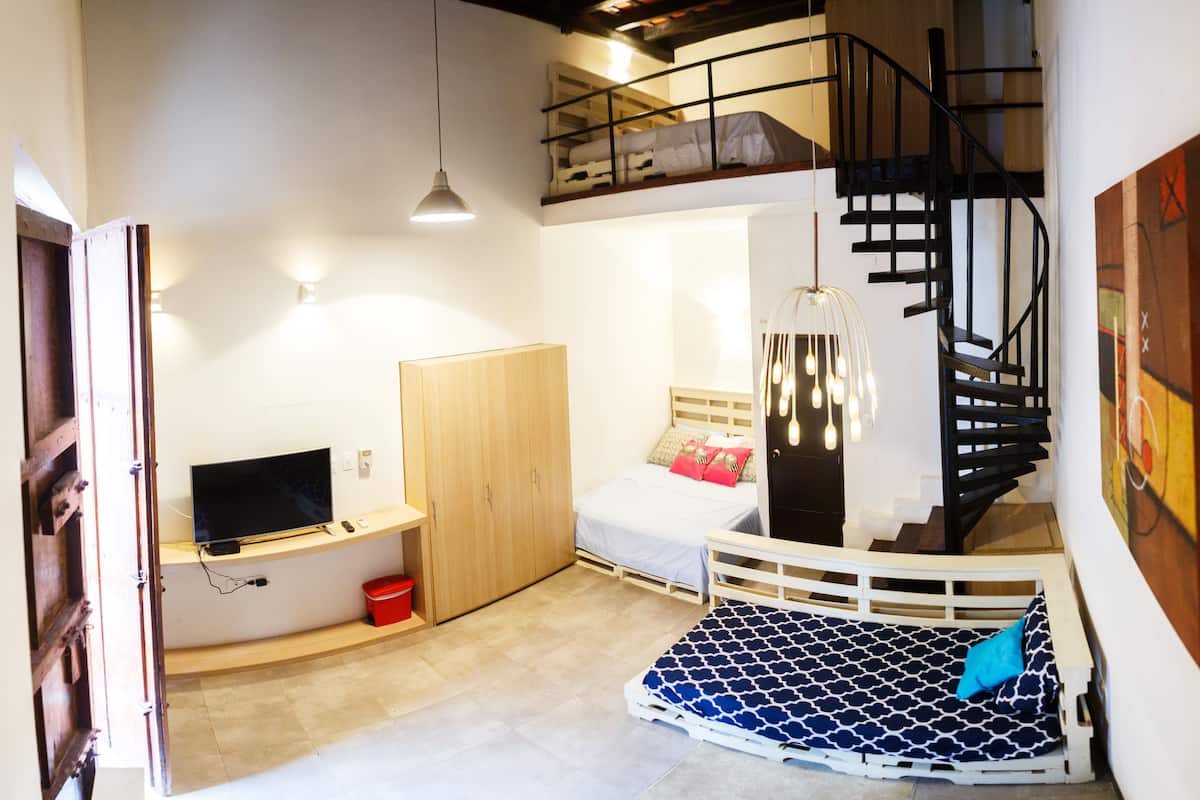 A compact studio apartment with a bunk bed, a desk with a computer, a wardrobe, a staircase, and a hanging light fixture.