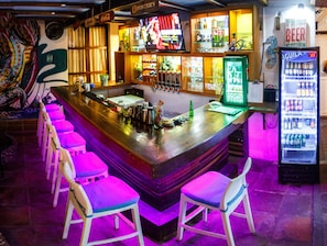 Een bar met een toonbank, krukken, een neonbord en een koelkast.
