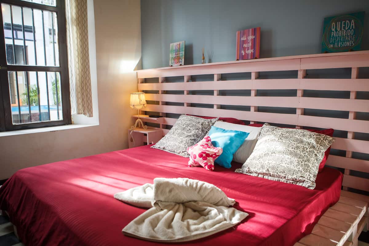 A neatly made bed with a red bedspread and pillows, a bedside lamp, and a window with bars.