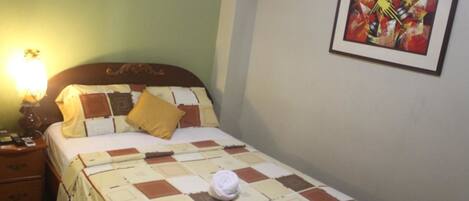Una cama bien hecha con una colcha a cuadros, una sola almohada y una toalla enrollada. Hay una mesita de noche con una lámpara y un teléfono. Un cuadro abstracto enmarcado está en la pared sobre la cama.