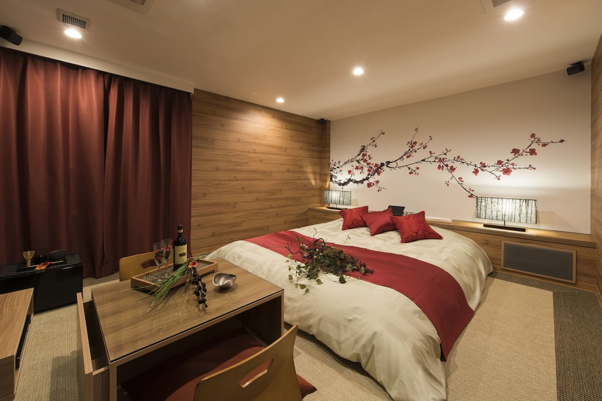 Un dormitorio moderno con cabecera de madera, una cama grande con ropa de cama roja, una mesita de noche con lámpara y un televisor montado en la pared.