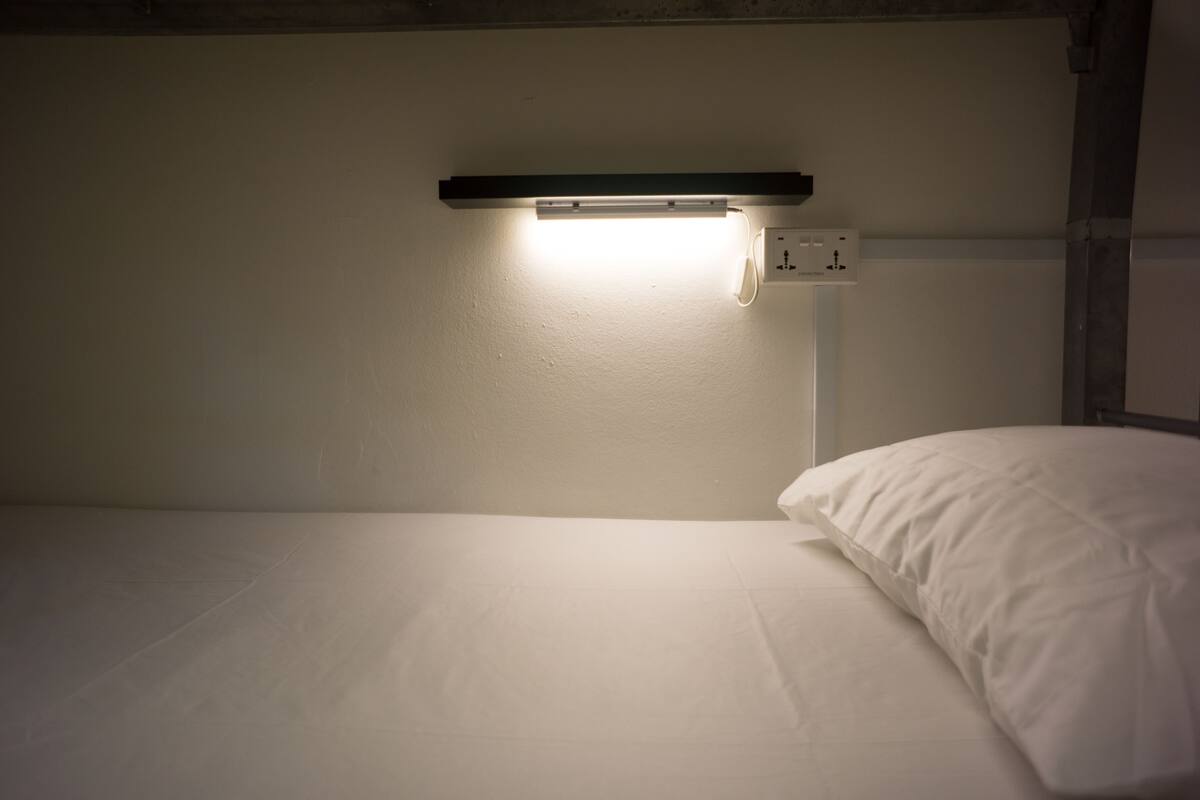 A bunk bed with a single white mattress and pillow, a bedside table with a lamp, and a wall socket.