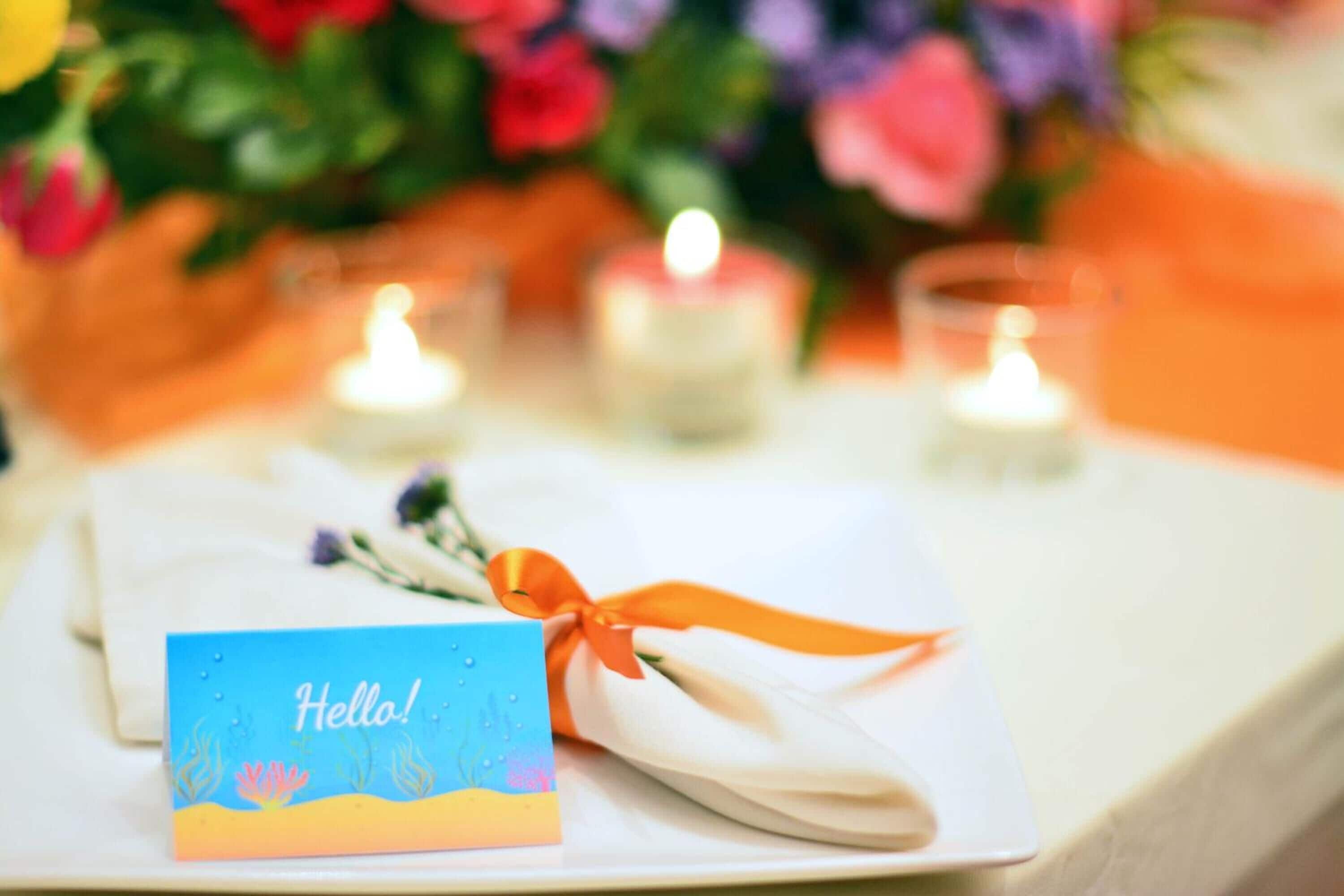 A table setting with a folded napkin tied with an orange ribbon, a card with 'Hello!' written on it, and three lit candles.