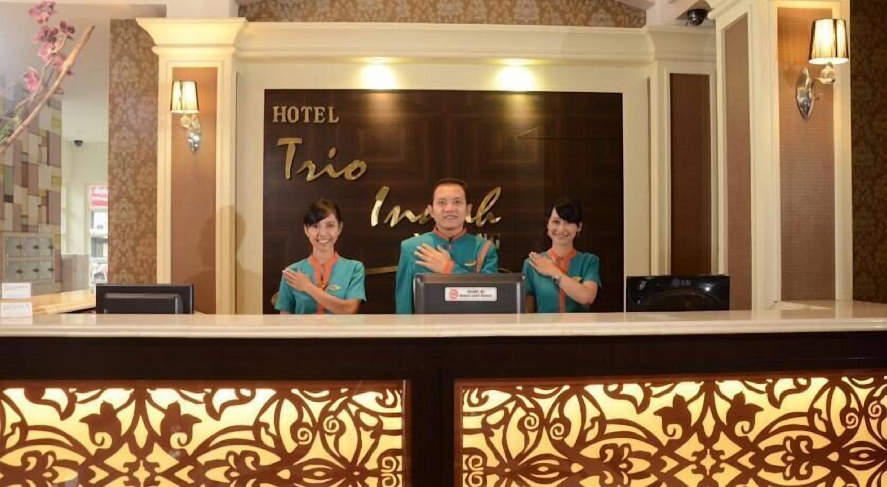 A hotel reception desk with three staff members, a sign reading "HOTEL Trico Inul", and decorative wall panels.