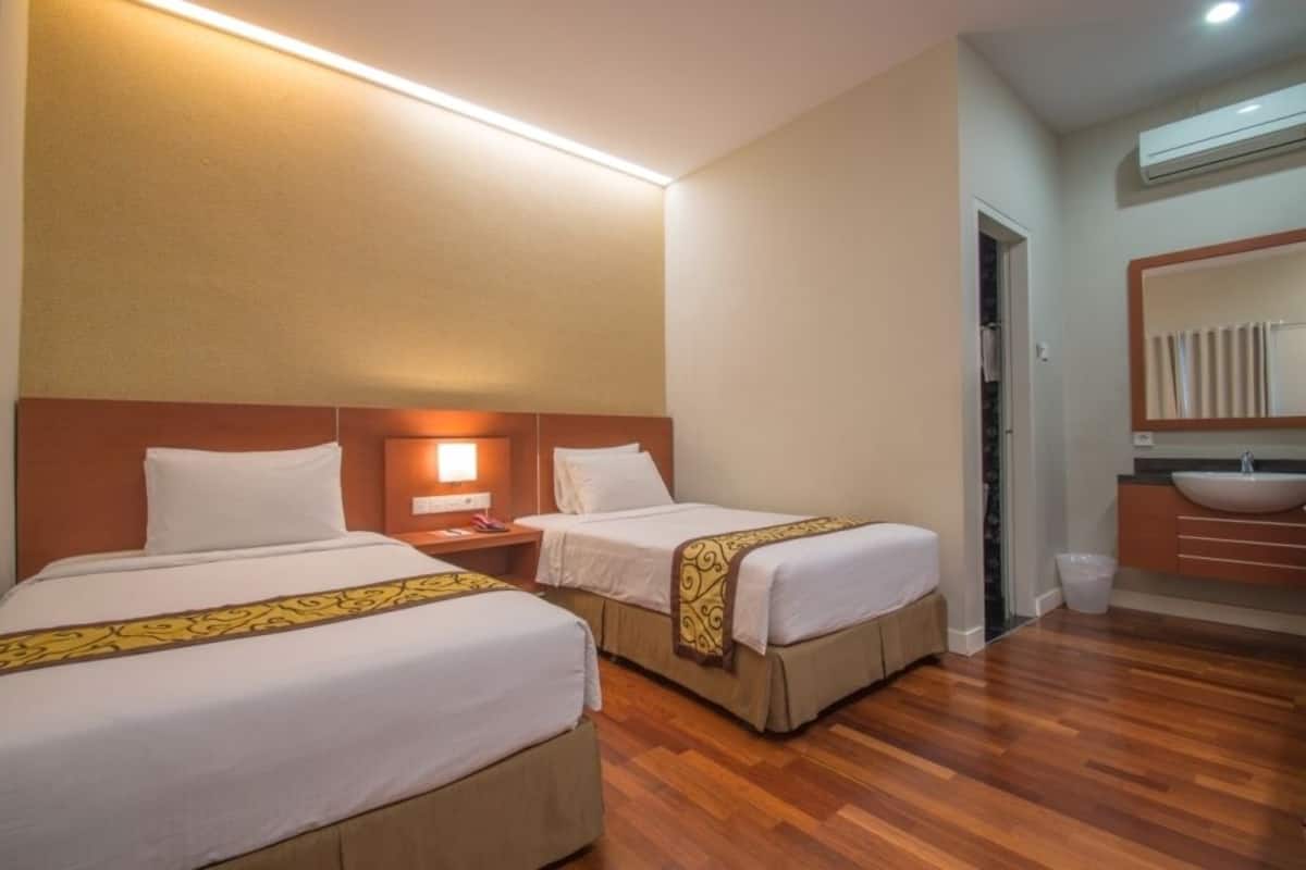 A hotel room with two beds, a nightstand, a bedside lamp, a bathroom with a sink and mirror, and wooden flooring.