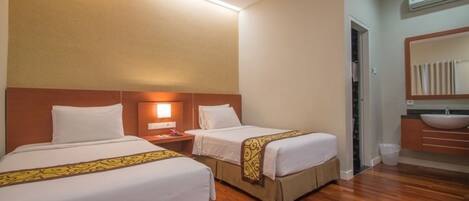 A hotel room with two beds, a nightstand, a bedside lamp, a bathroom with a sink and mirror, and wooden flooring.