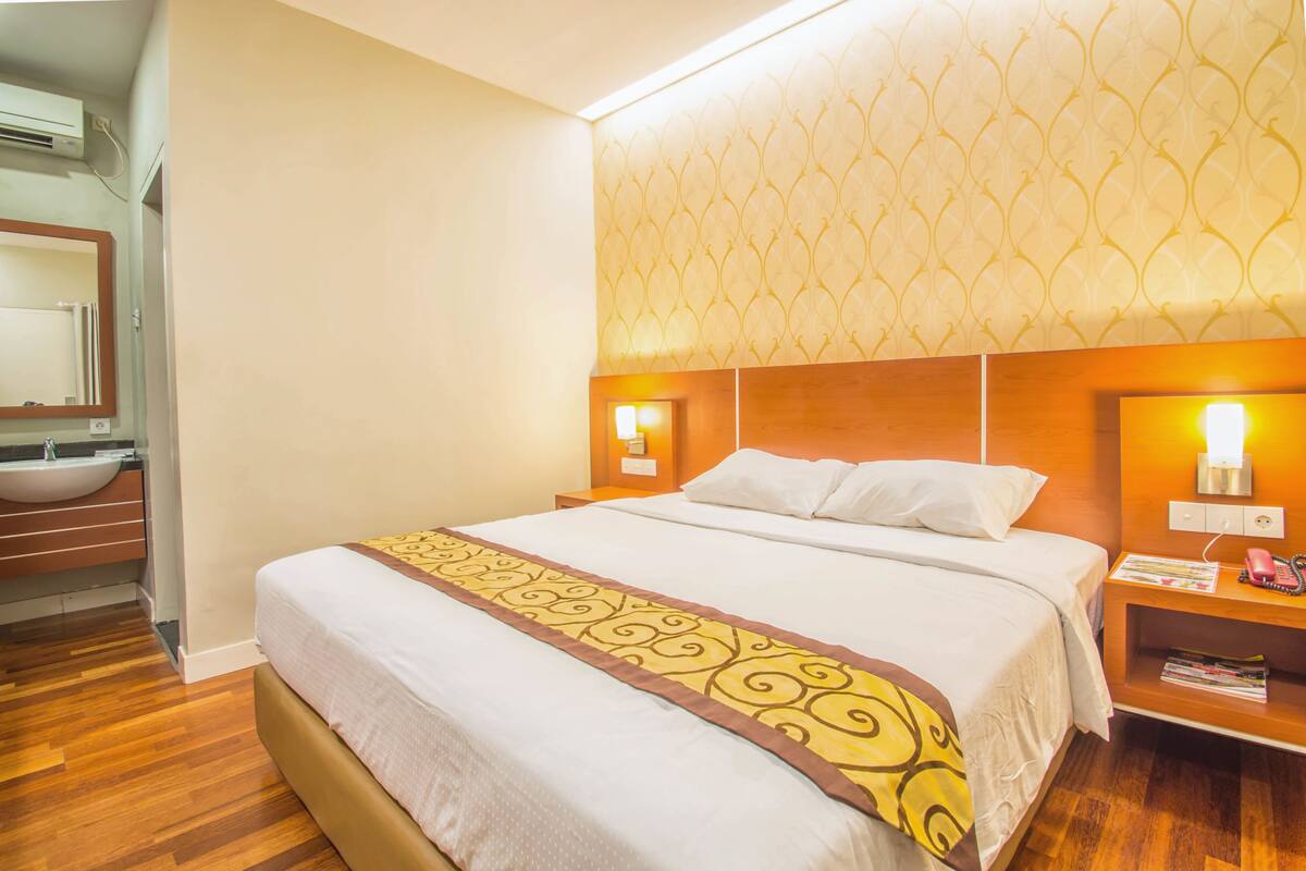 A hotel room with a large bed, a bedside table with a lamp, a nightstand with a phone, and a bathroom with a sink and mirror.