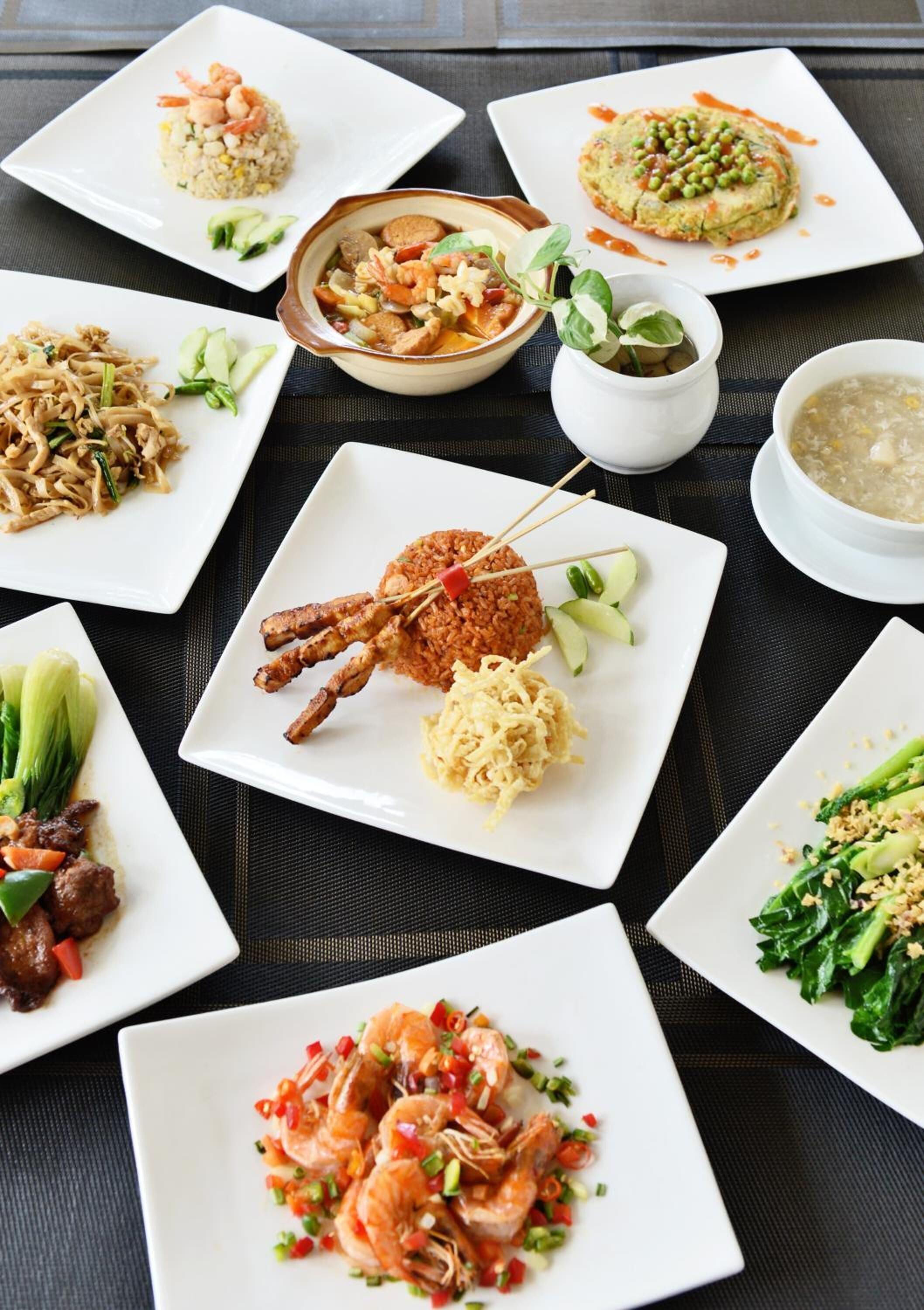Une variété de plats présentés sur des assiettes carrées blanches, notamment des crevettes, des nouilles et un plat de légumes, disposés sur une nappe sombre.
