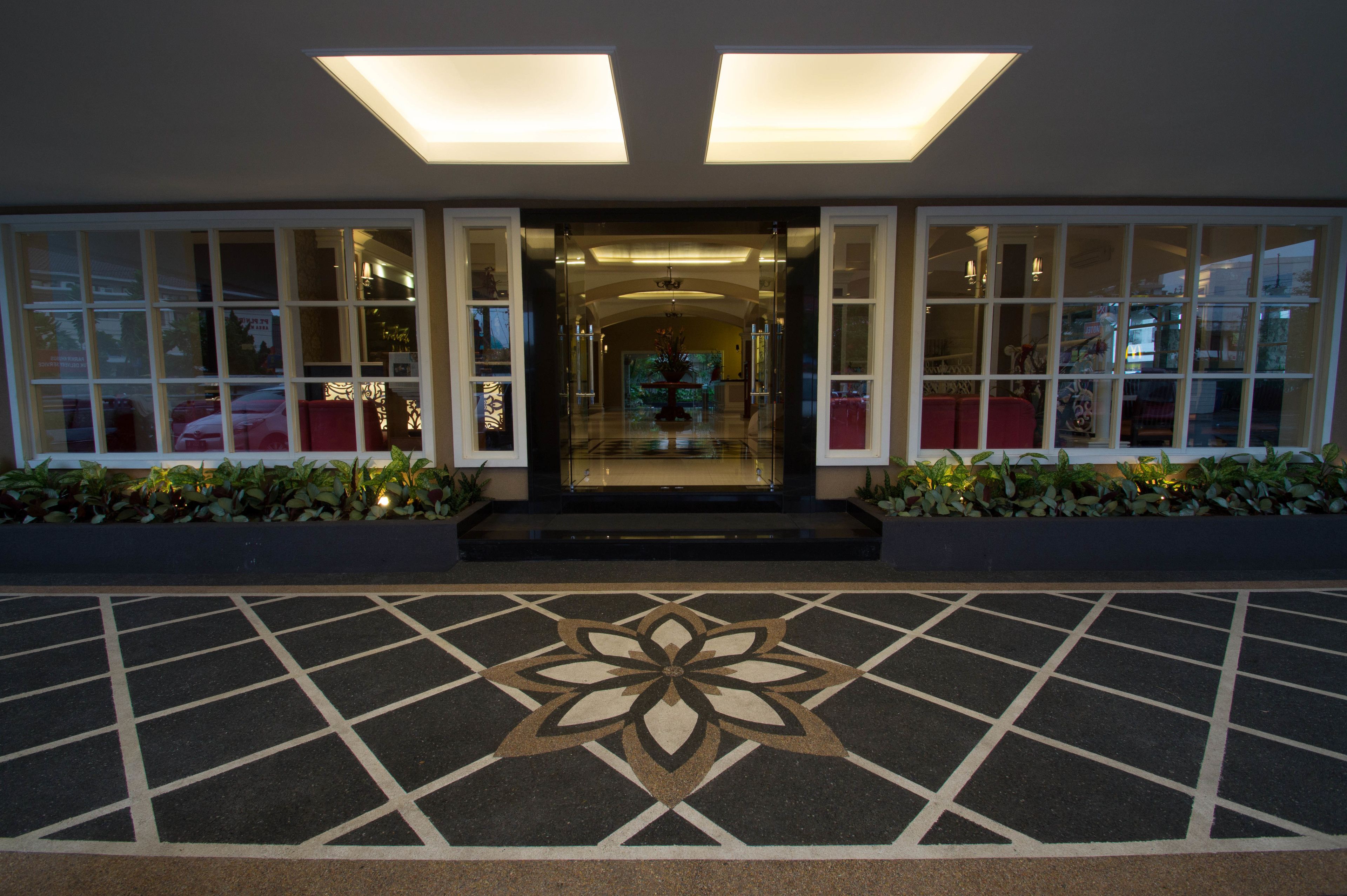 A hotel entrance with a geometric floor design, a central floral pattern, and a glass door leading to a lobby with seating and a chandelier.