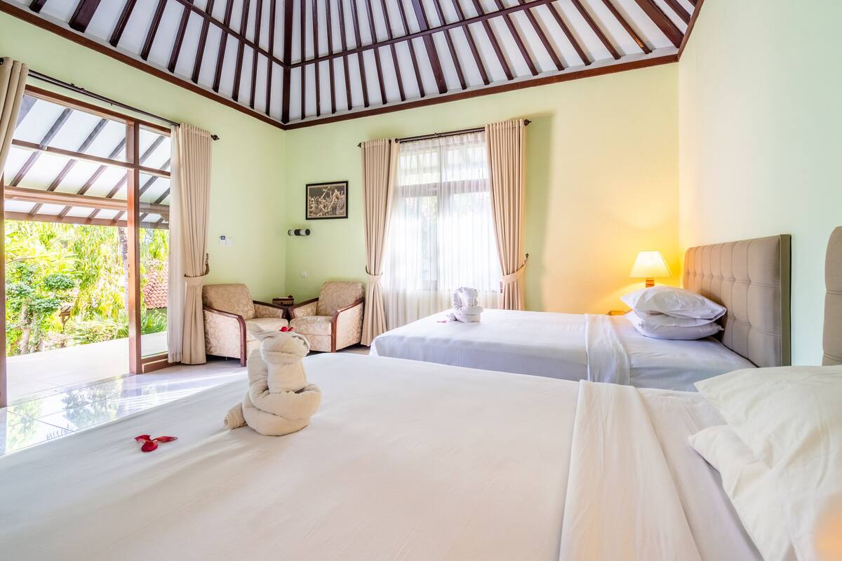 A hotel room with two beds, a sitting area with a chair, a table, and a view of greenery outside.