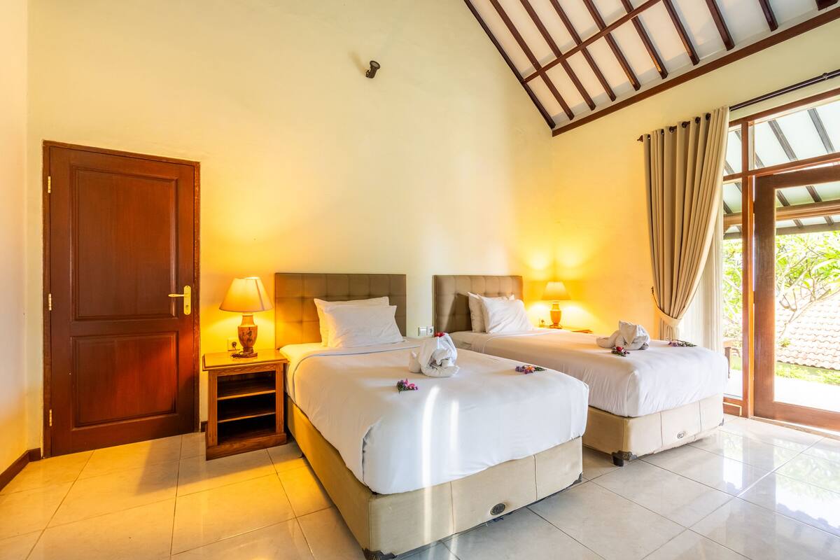 A hotel room with two beds, a wooden door, a nightstand with a lamp, and a view of greenery through the window.