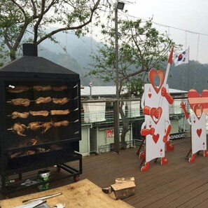 Un barbecue avec plusieurs brochettes de viande, une terrasse en planches de bois et, en arrière-plan, des arbres et des montagnes.