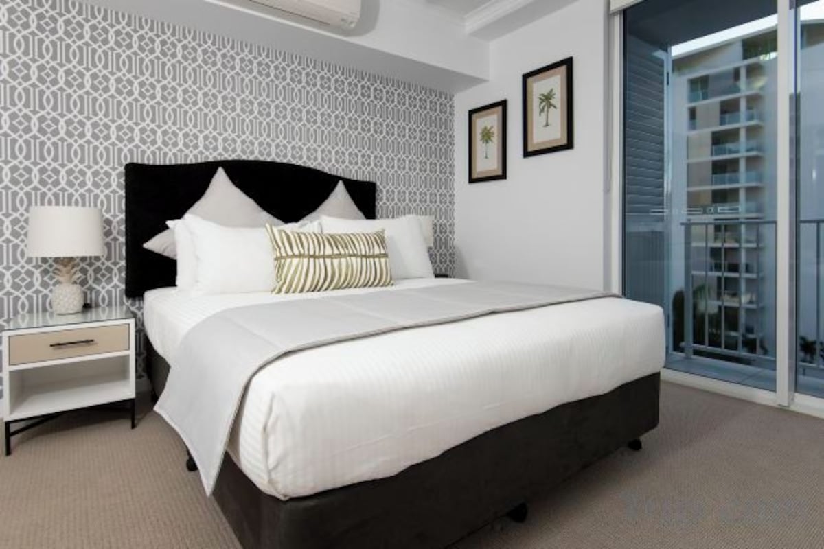 A hotel room with a large bed, a nightstand, a lamp, and a view of a balcony.