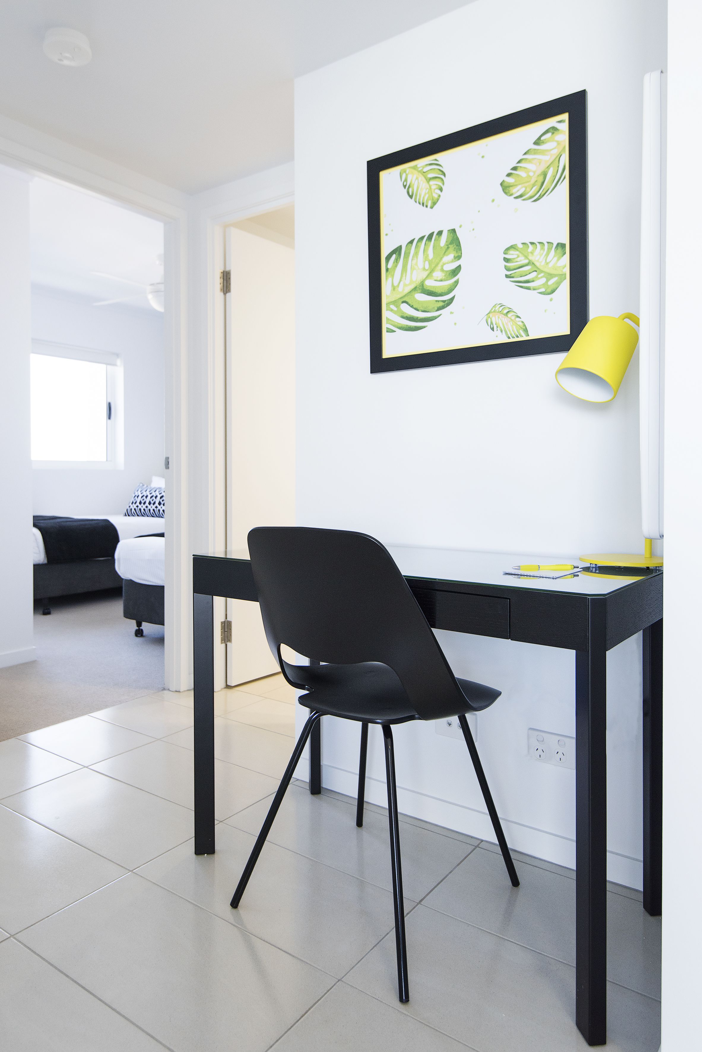 A modern room with a black desk, a black chair, a yellow lamp, and a framed leaf pattern on the wall.