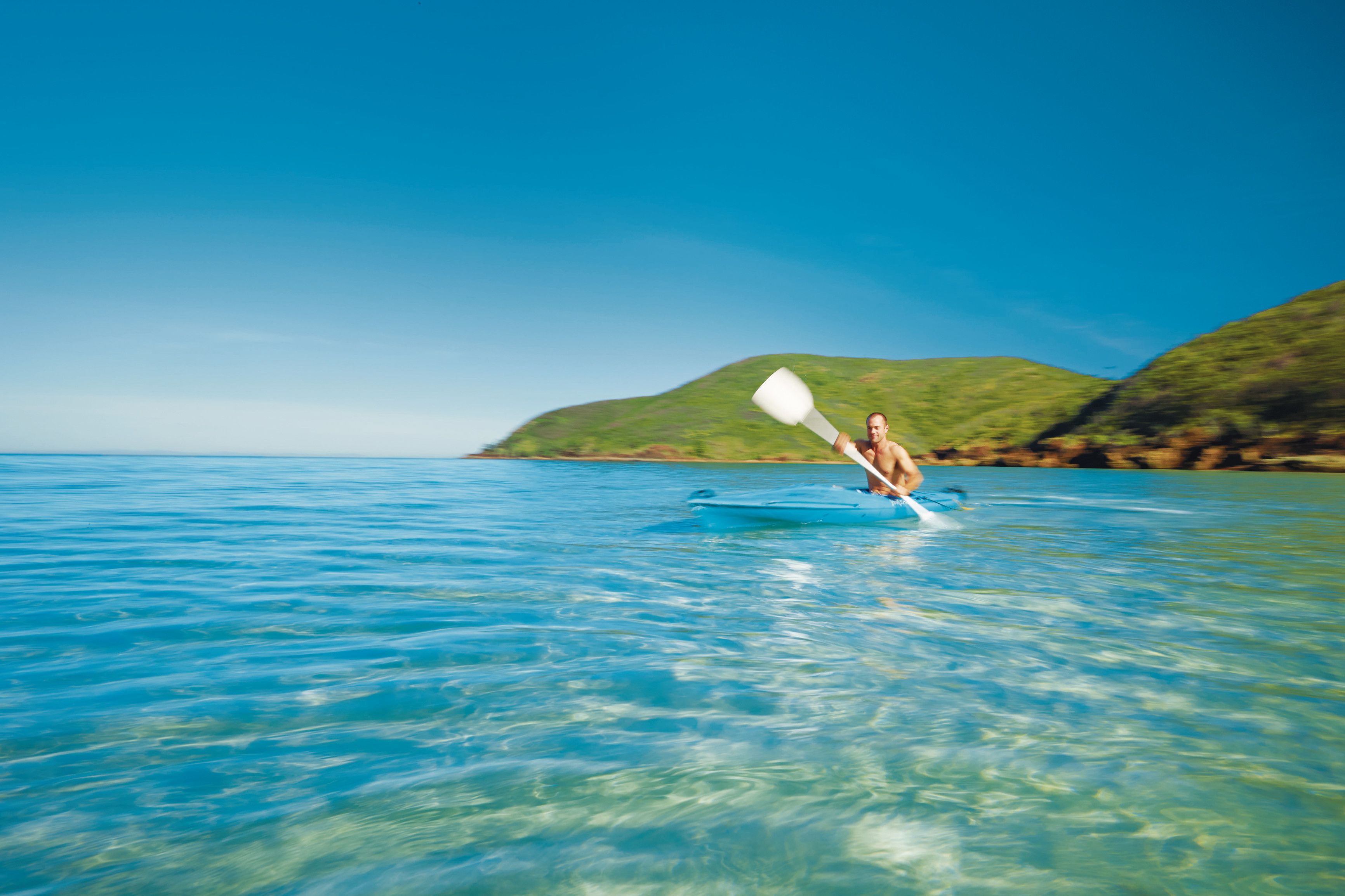 A person kayaking on clear blue water with a hilly landscape in the background.