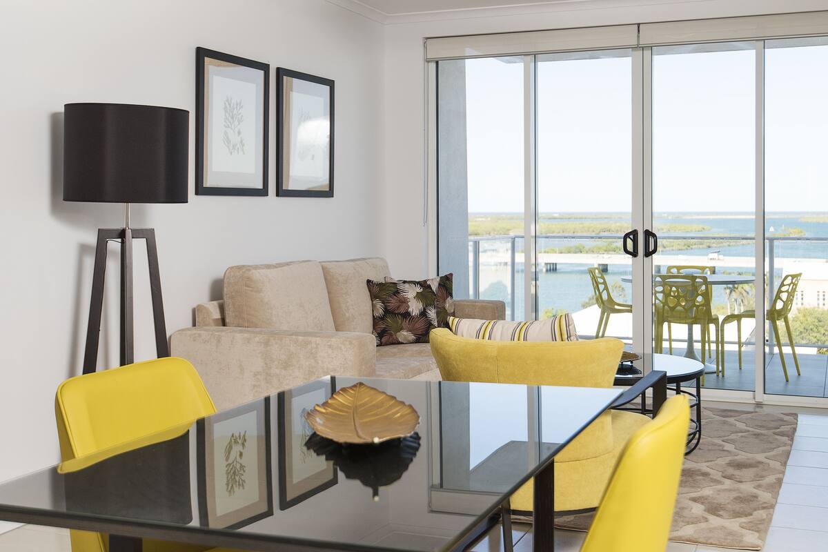 A modern living room with a sofa, yellow chairs, a glass table, and a balcony with a view of the sea.