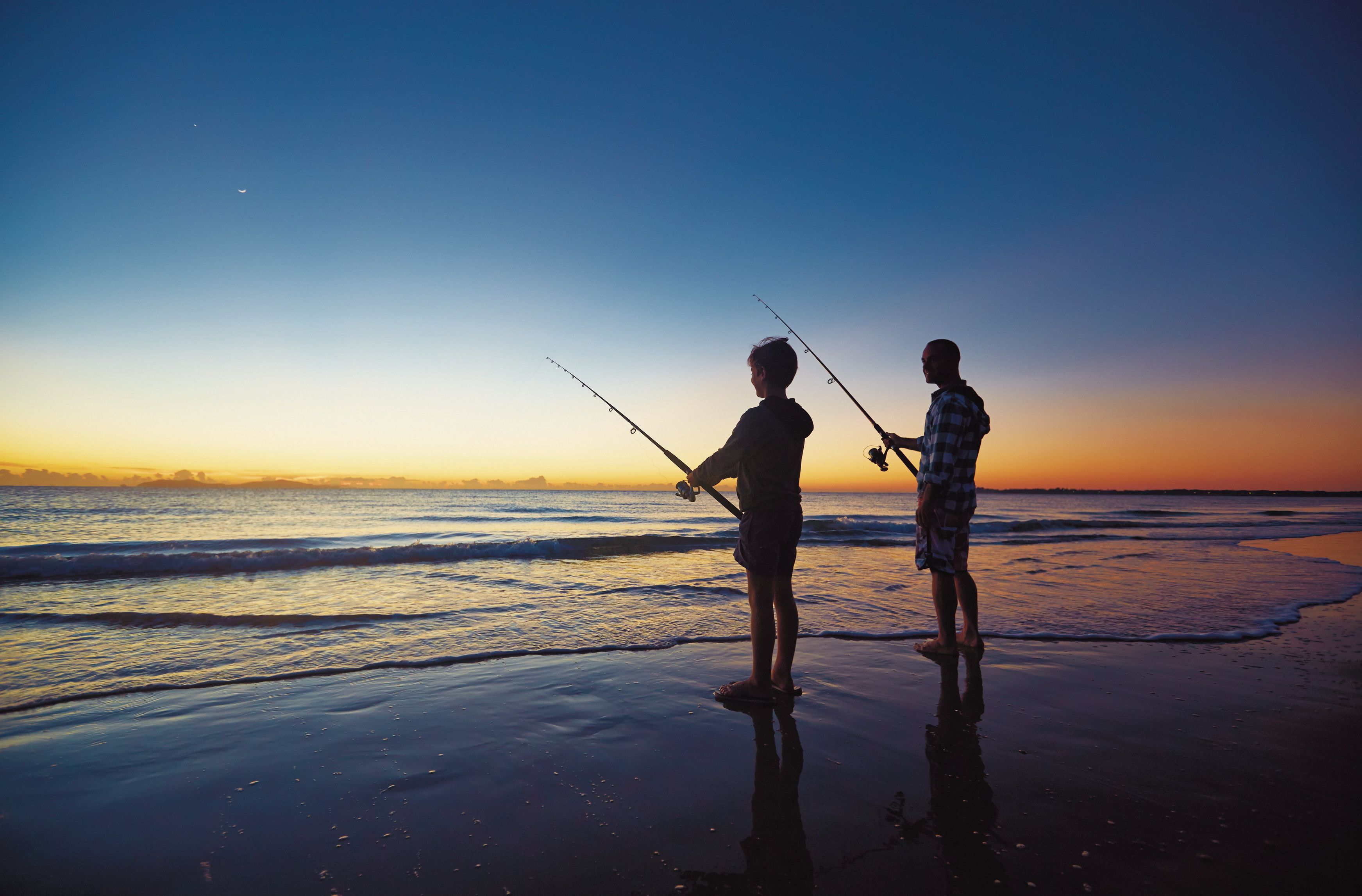 Two people fishing on a beach at sunset.