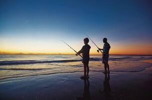 Two people fishing on a beach at sunset.