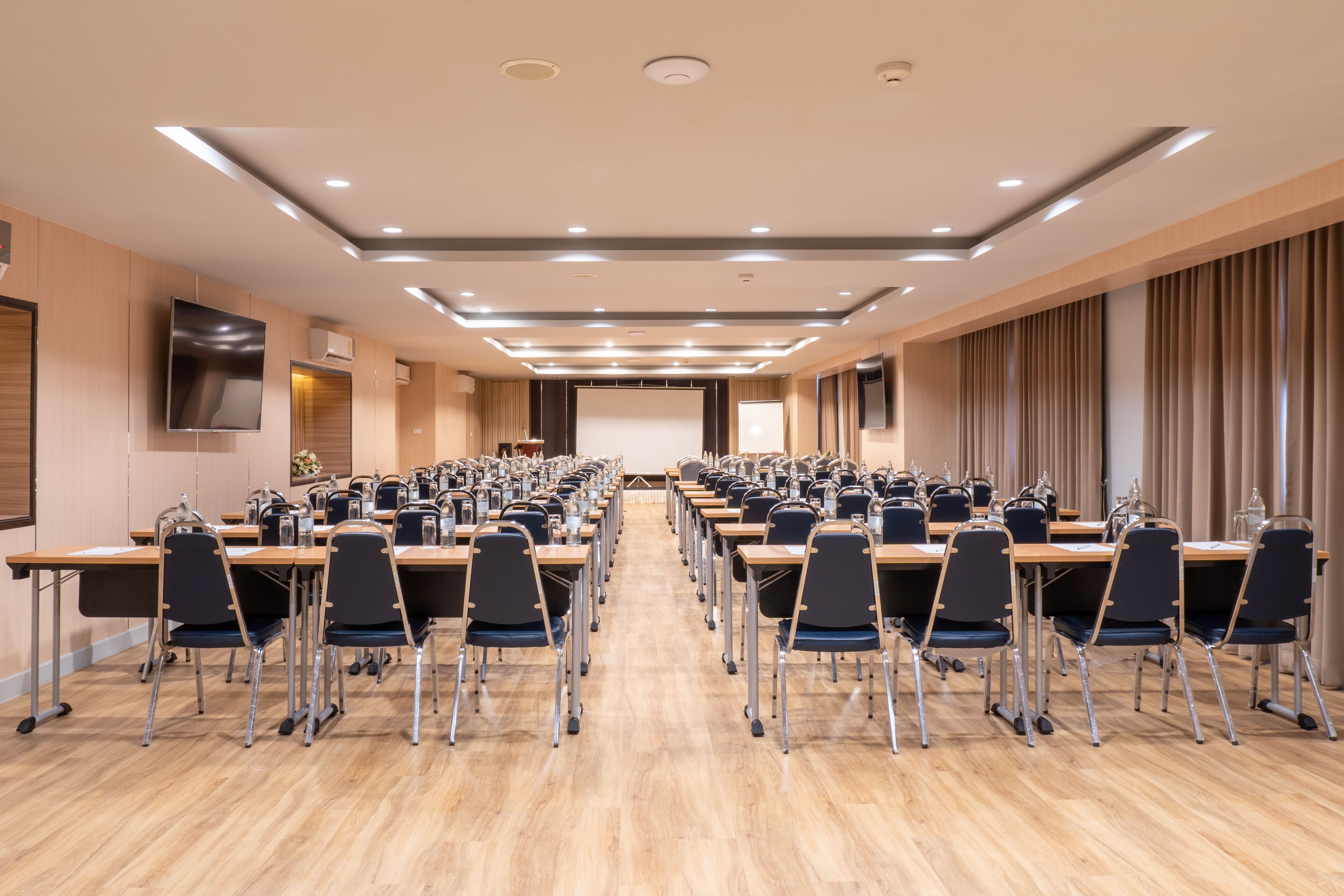 Une salle de conférence avec des rangées de tables et de chaises, un grand écran et un pupitre.