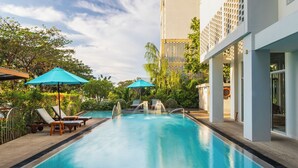 Una piscina con sillas de descanso y sombrillas, rodeada de vegetación y un edificio moderno.
