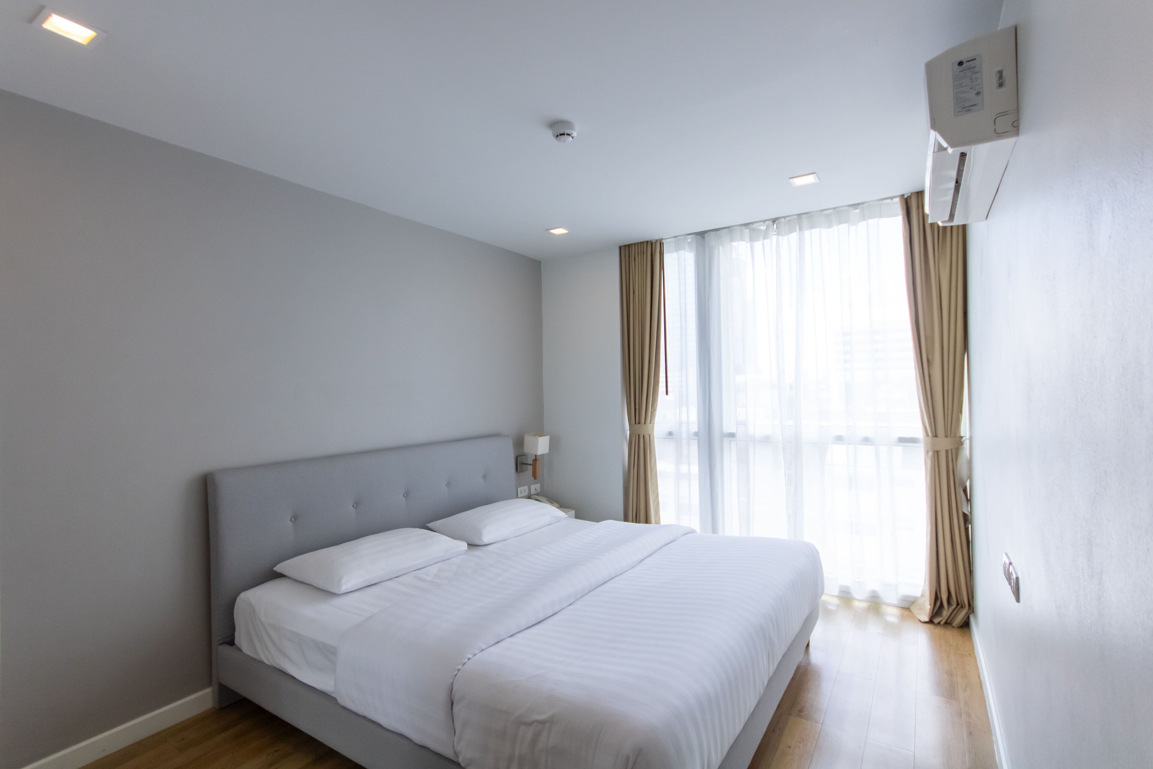 A modern hotel room with a large bed, a nightstand, a wall-mounted air conditioner, and a window with curtains.