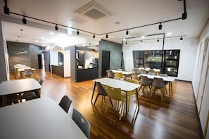Un comedor moderno con varias mesas y sillas, una cocina al fondo y ventanas amplias.