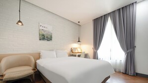 Een hotelkamer met een groot bed, een stoel, een nachtkastje, een lamp, een afbeelding van Parijs en een raam met gordijnen.