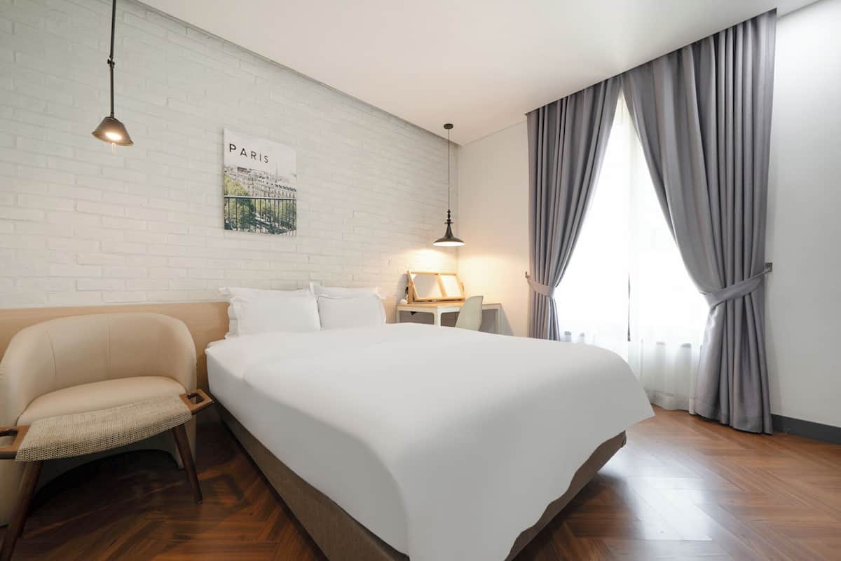 Ein Hotelzimmer mit einem großen Bett, einem Sessel, einem Nachttisch, einer Lampe, einem Bild von Paris und einem Fenster mit Vorhängen.