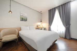 Une chambre d’hôtel avec un grand lit, une chaise, une table de chevet, une lampe, un tableau représentant Paris et une fenêtre avec des rideaux.