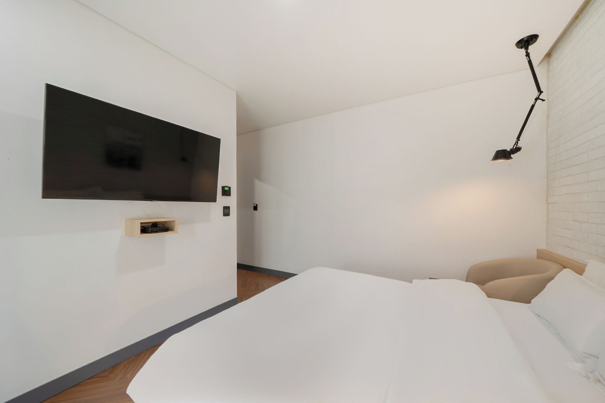 Un dormitorio moderno con una televisión de pantalla plana montada en la pared, una mesita de noche, una cama con ropa de cama blanca y una lámpara de escritorio negra.