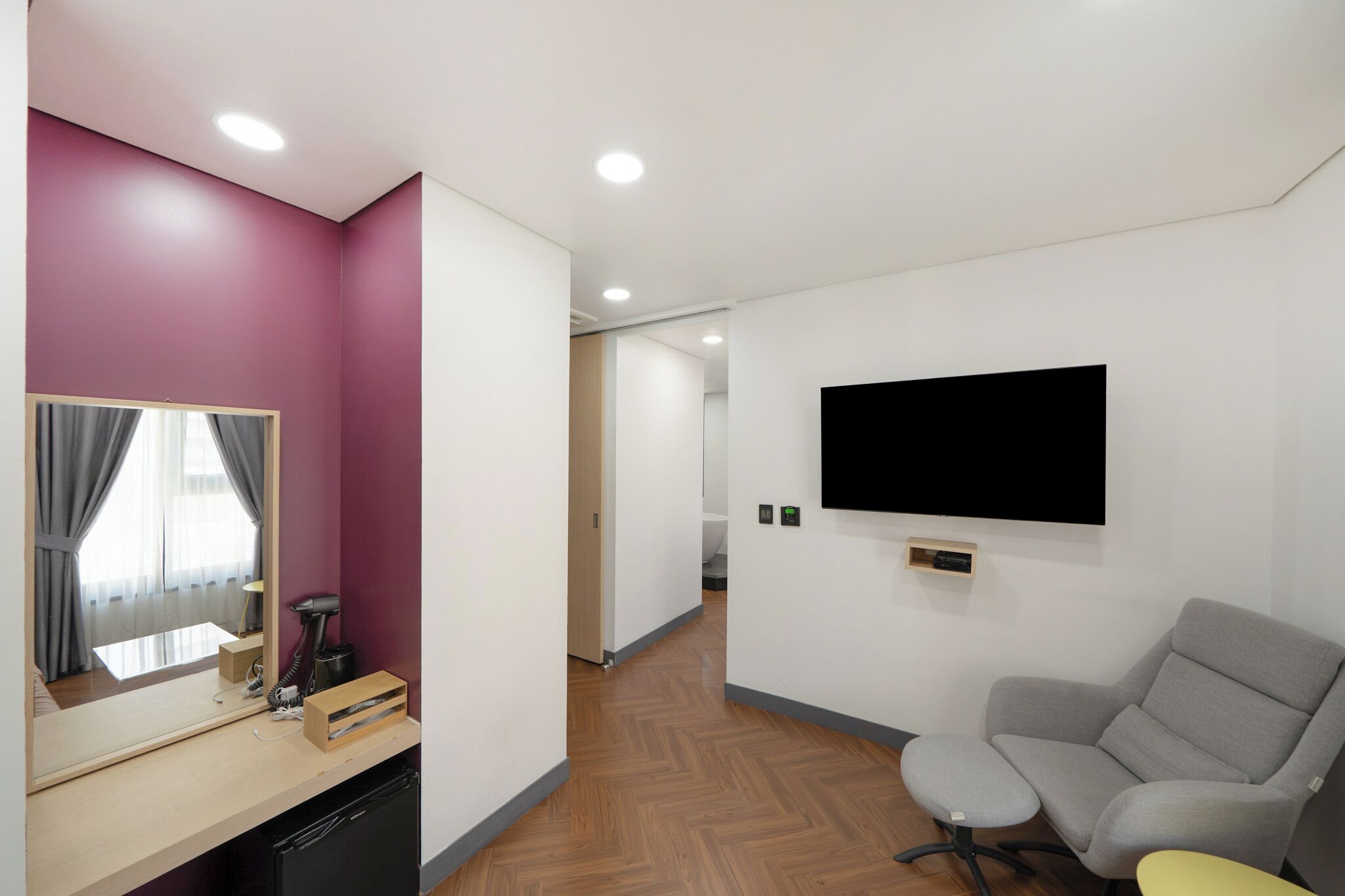 A hotel room with a wooden floor, a mirror, a hairdryer, a TV mounted on the wall, and a grey armchair.