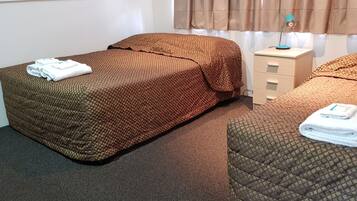 A hotel room with two beds, each with a brown patterned bedspread and white towels. There is a small bedside table with a lamp and a drawer unit.