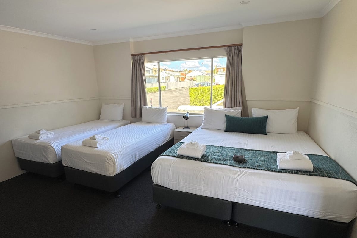 Habitación de hotel con dos camas individuales, un ventanal con cortinas, una mesita pequeña y suelo alfombrado.