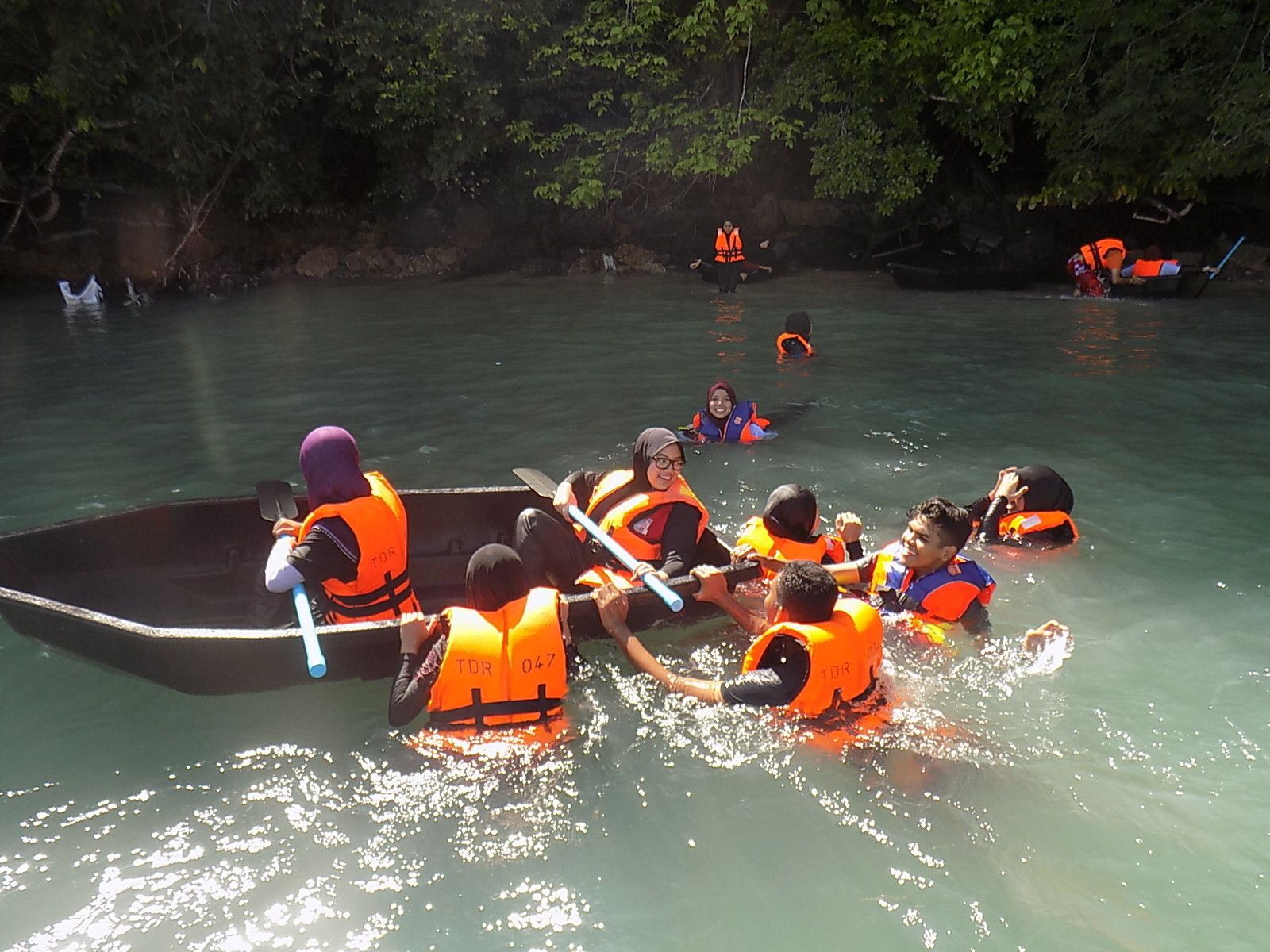 Un grupo de personas en un bote, con chalecos salvavidas, remando en un río tranquilo rodeado de árboles.