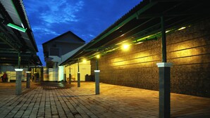 A covered outdoor area with a tiled floor, a brick wall, and a building with a sloped roof in the background. - The Ayudya Glogor Carik (Denpasar)