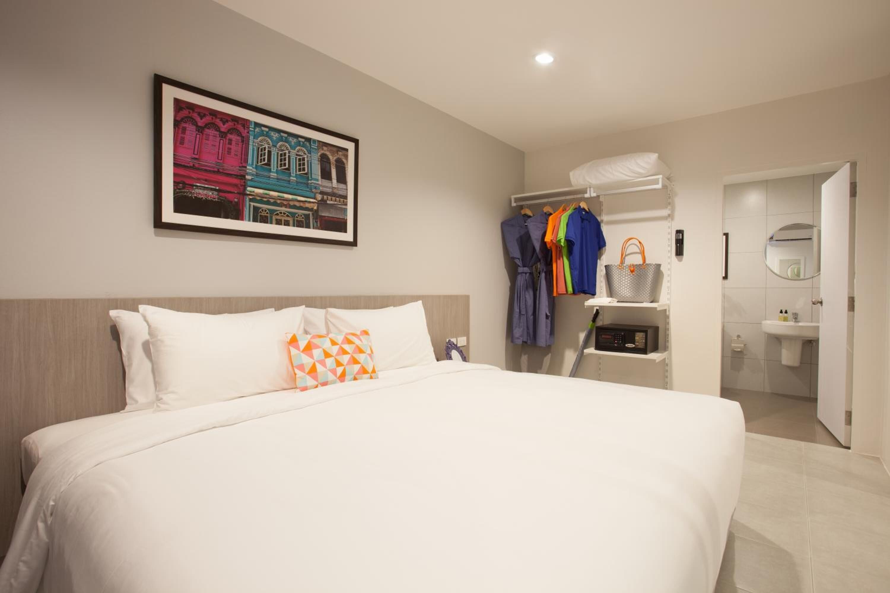 A hotel room with a large bed, a framed picture on the wall, a wardrobe with hanging clothes, and a bathroom visible through an open door.