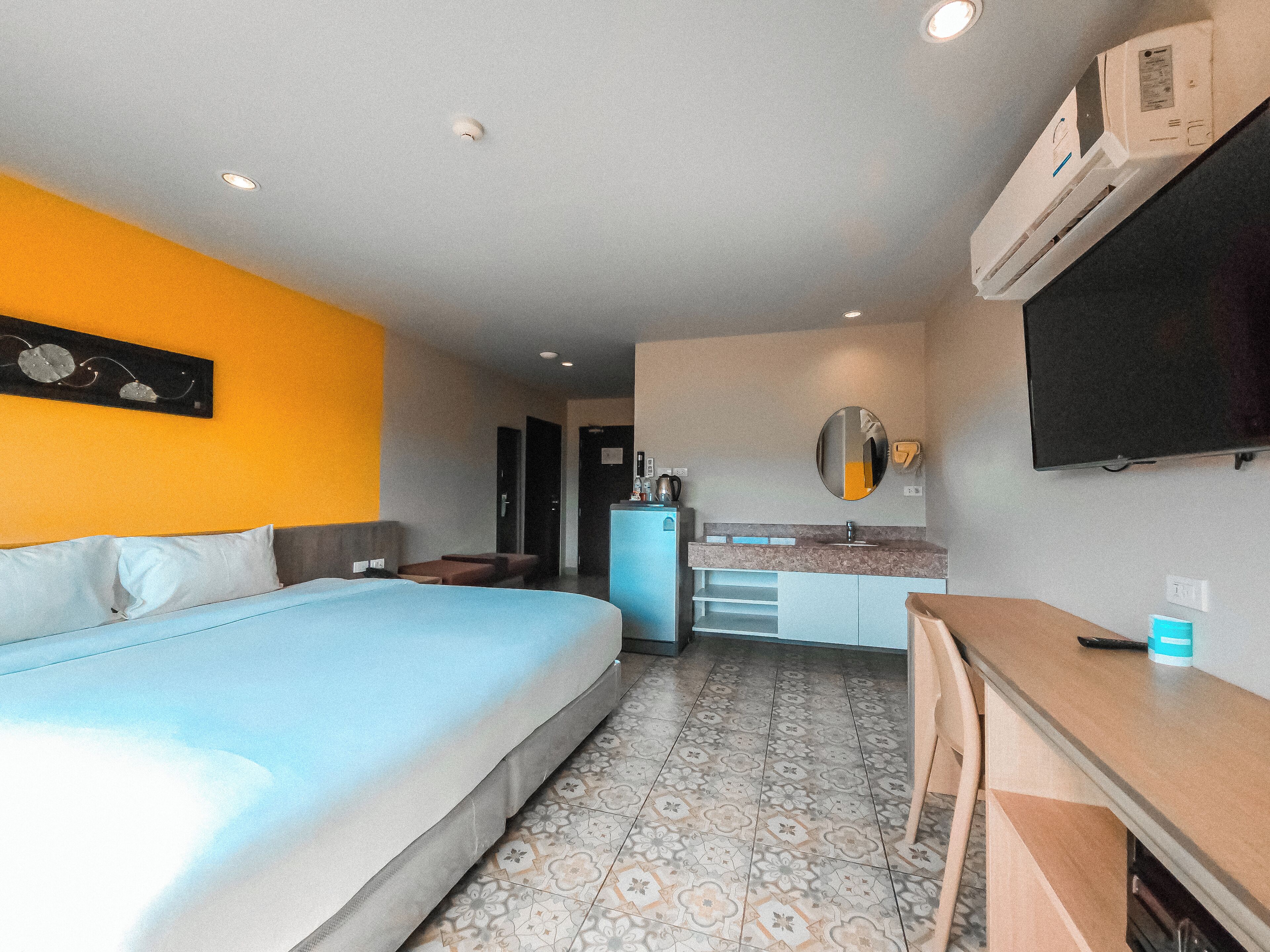 A hotel room with a bed, a desk, a small kitchenette, a TV, and a bathroom visible through an open door.