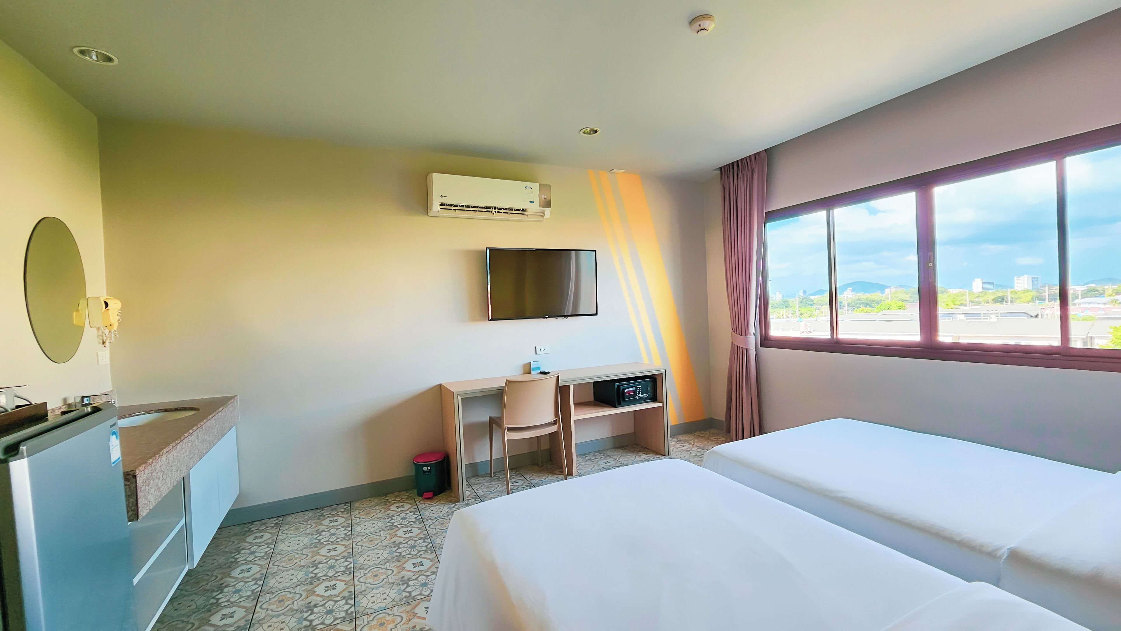 A hotel room with a bed, a desk with a chair, a TV mounted on the wall, and a window with a view of the city.