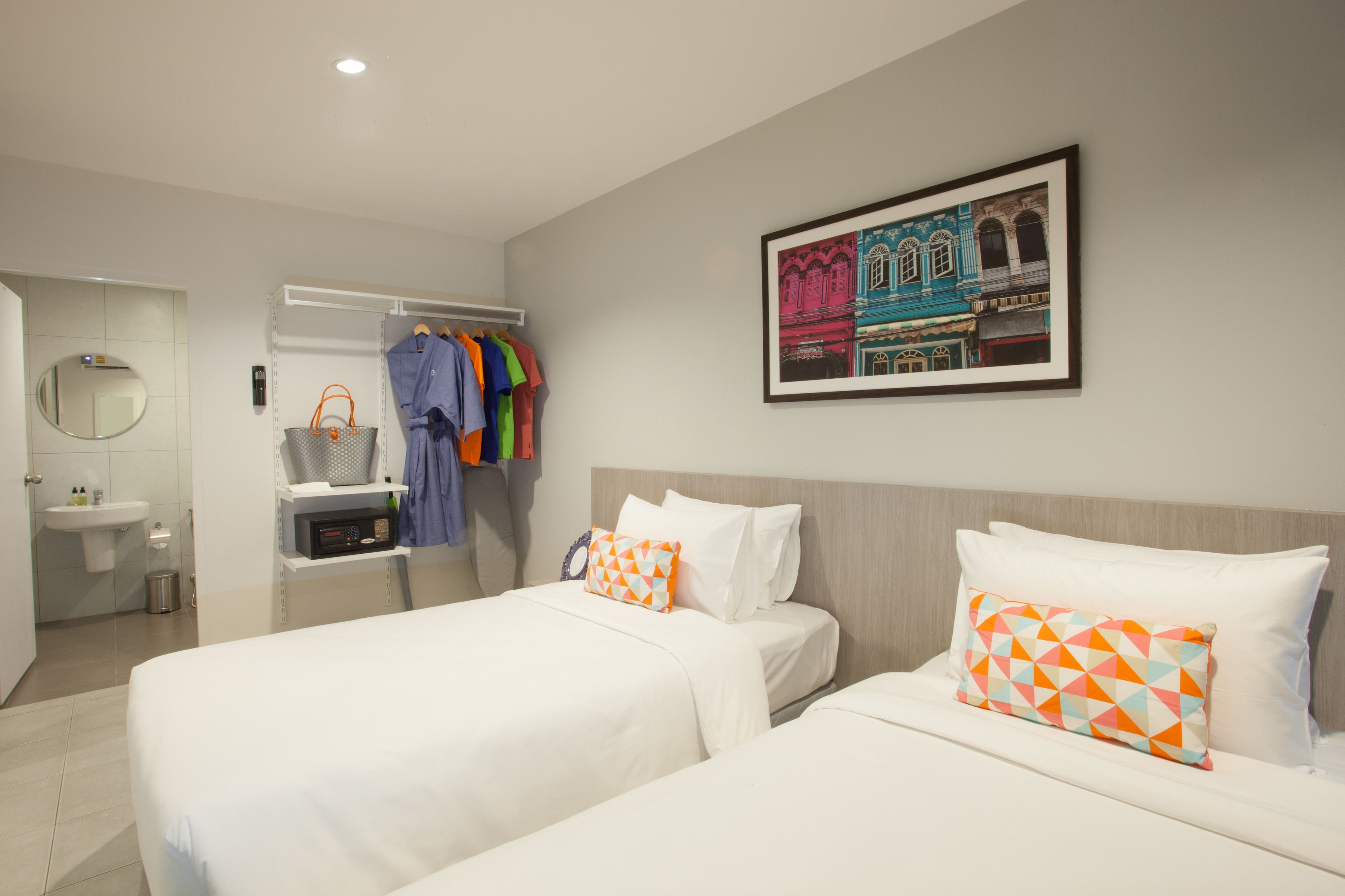 A hotel room with two beds, a bathroom, a coat rack with colorful clothes, and a framed picture on the wall.