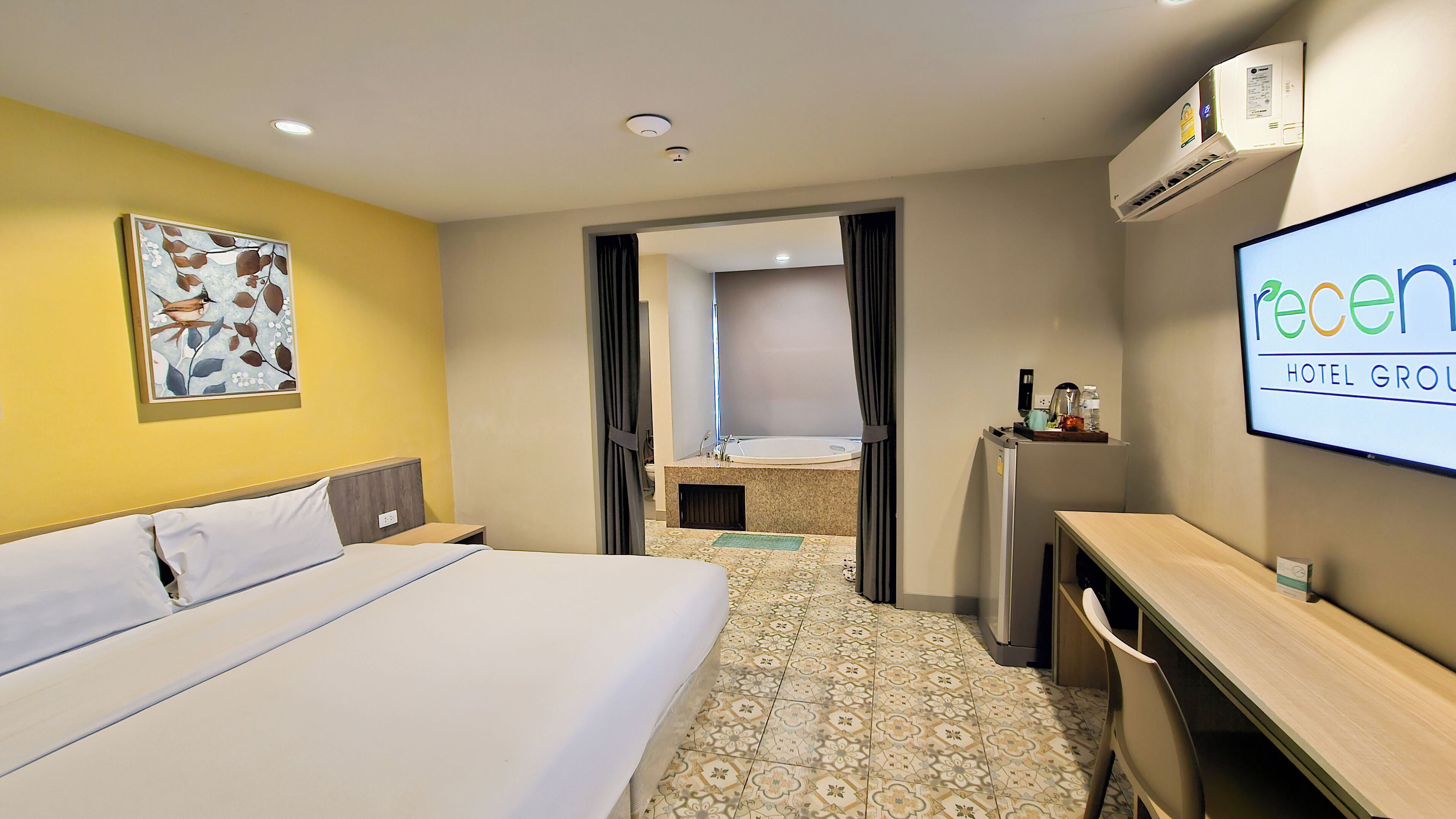 A hotel room with a bed, a desk, a TV displaying the hotel name, and a bathroom visible through an open door.
