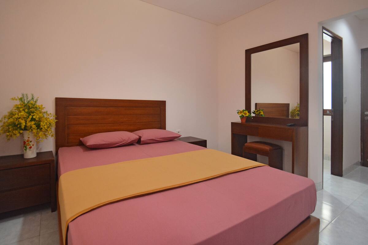 A bedroom with a bed, bedside table, dresser, mirror, and a vase with flowers.