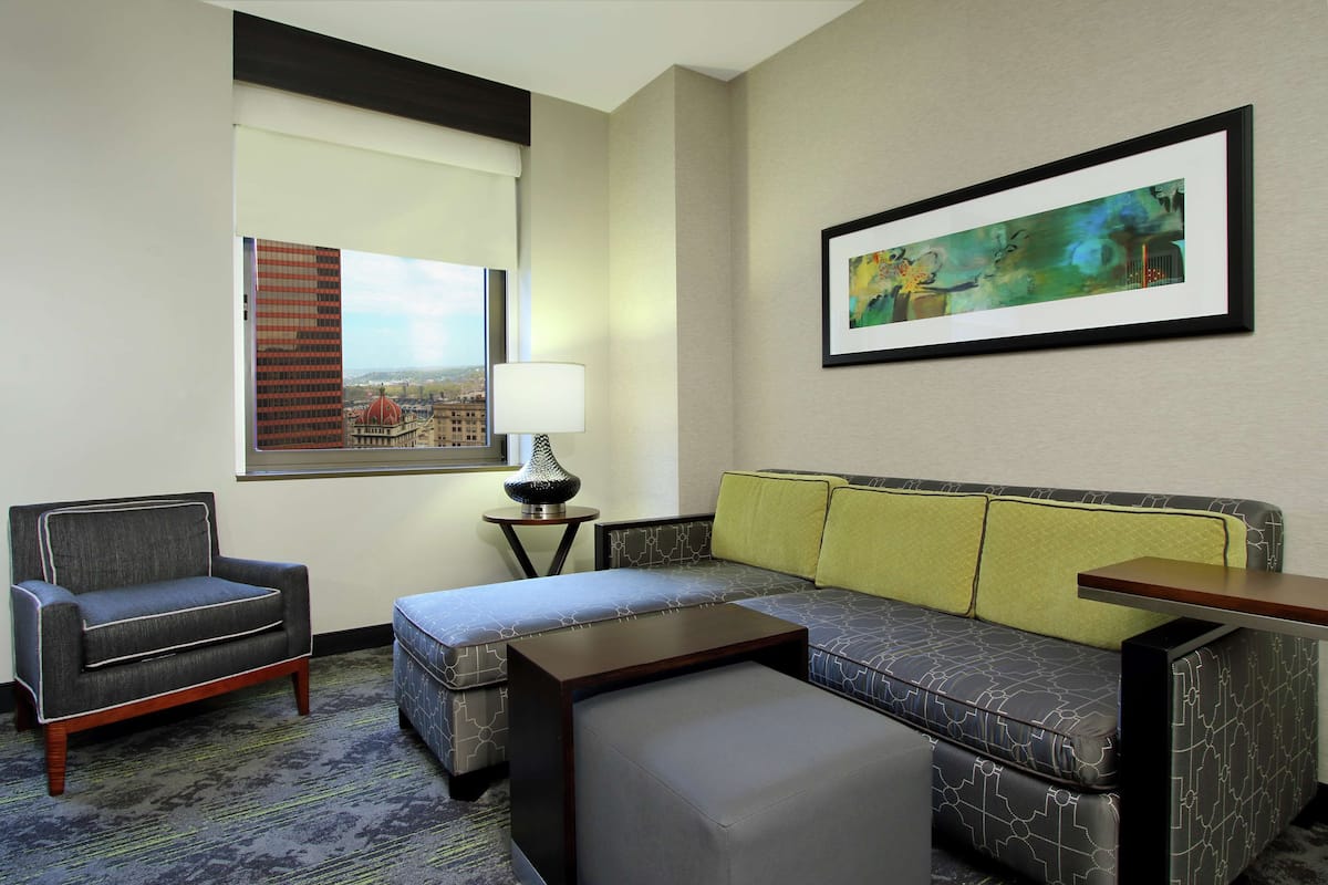 Una habitación de hotel con sofá, sillón, reposapiés y una mesa.