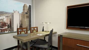A hotel room with a city view, a desk with a lamp, a chair, and a TV on the wall.