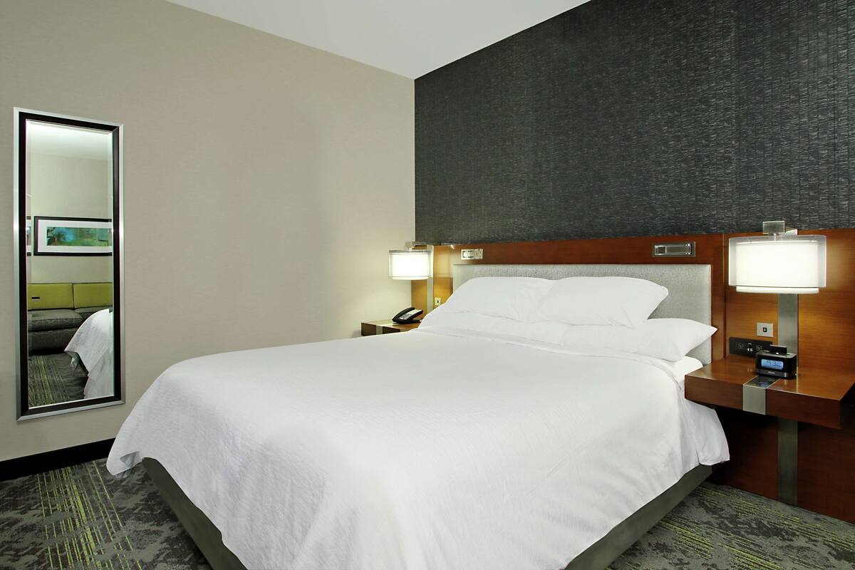 Una habitación de hotel con una cama grande, dos mesitas de noche con lámparas, un espejo y un reloj en la pared.