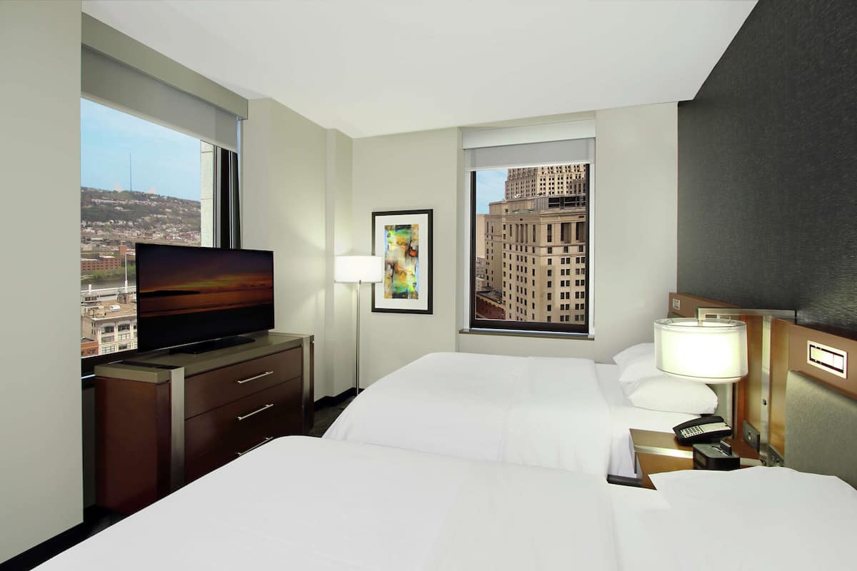 Habitación de hotel con cama, televisión, escritorio con lámpara y ventana con vista a la ciudad.