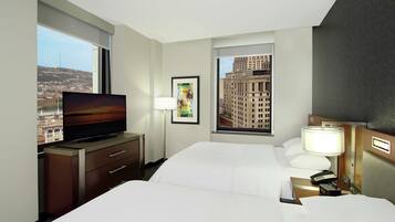 Quarto de hotel com cama, televisão, escrivaninha com abajur e janela com vista para a cidade.
