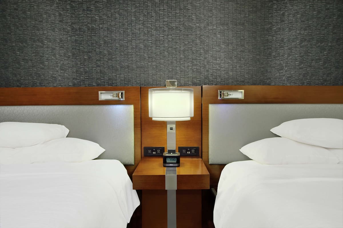 Habitación de hotel con dos camas, cabecera de madera, mesita de noche con un reloj y pared gris con textura.