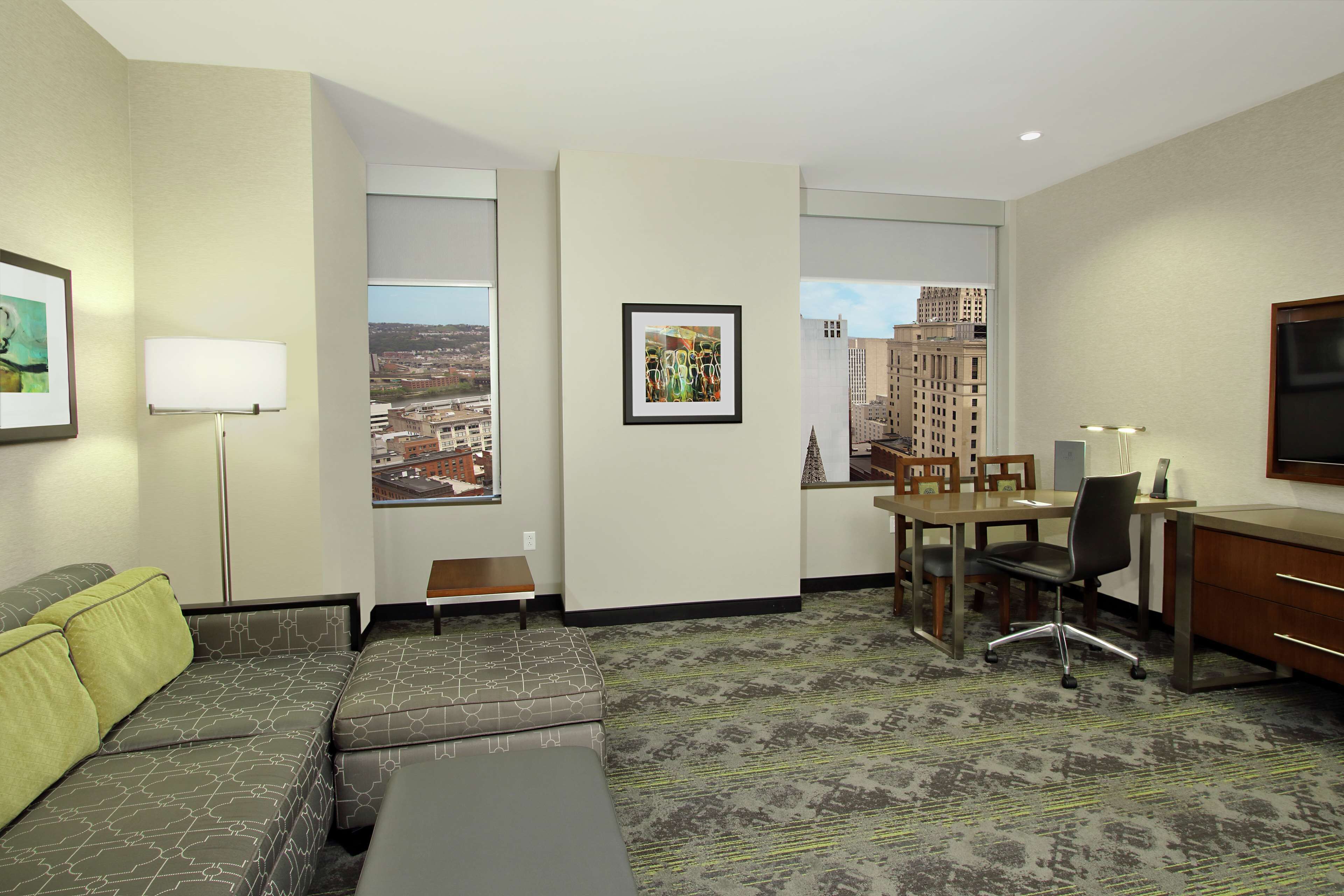 A hotel room with a sofa, a desk, a chair, a lamp, and a large window offering a city view.