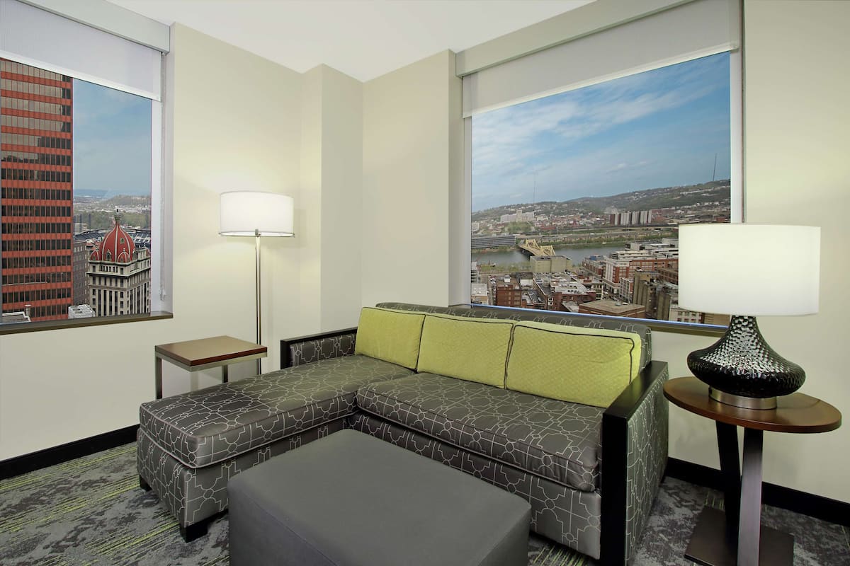 Habitación de hotel con sofá modular, una mesita, lámpara y ventanas amplias con vistas a la ciudad.