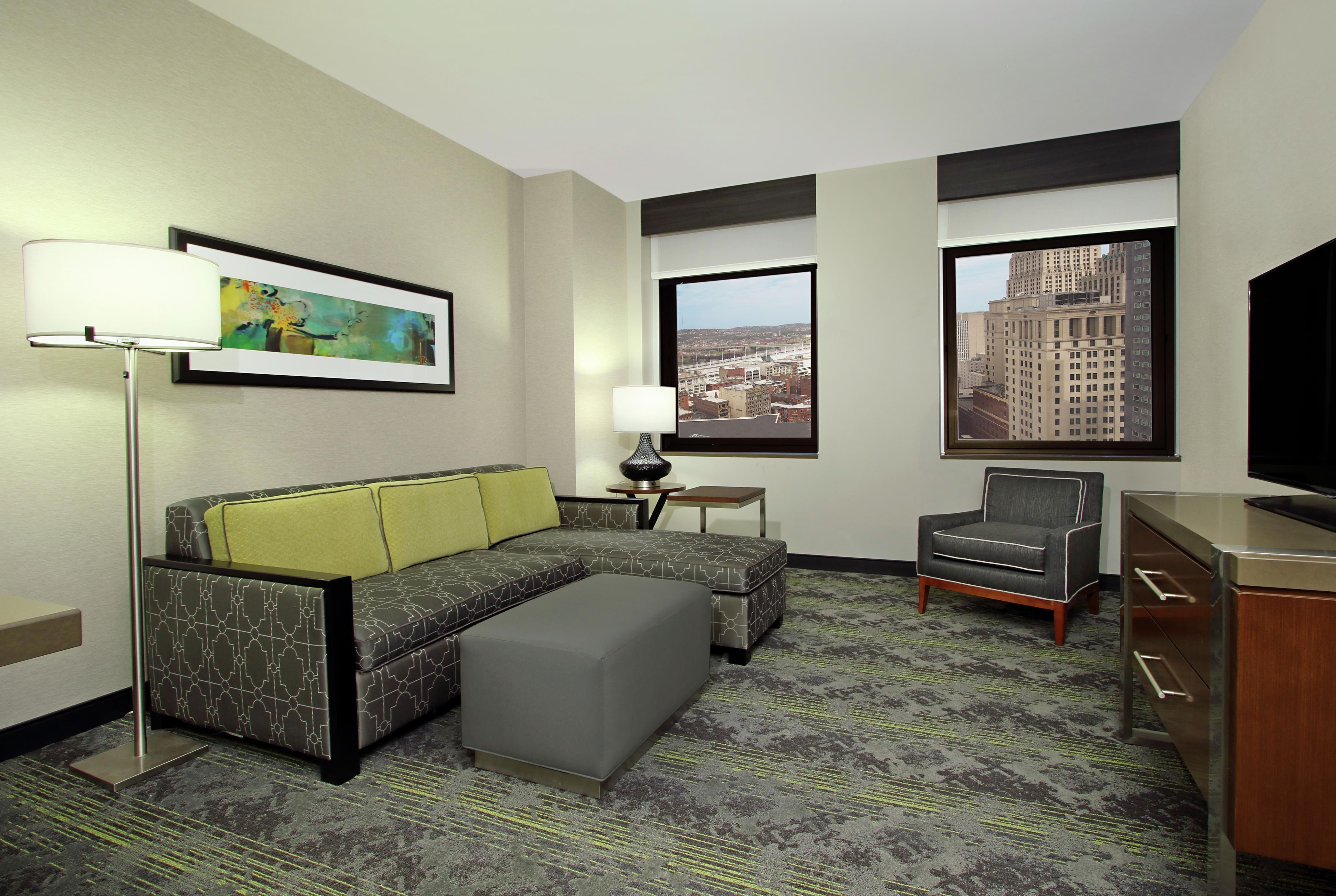 Habitación de hotel con sofá, reposapiés, sillón, televisor de pantalla plana, un cuadro y ventana con vista a la ciudad.